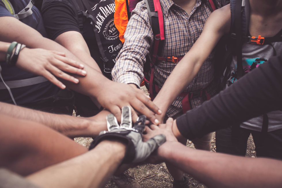 Group of diverse people with backpacks join hands in unity. Casual clothing, plaid shirt, black glove visible, outdoor setting, teamwork mood.
