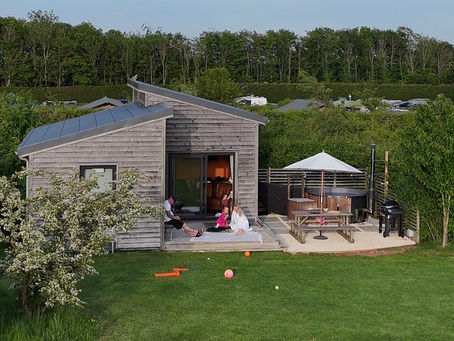 Family relaxing on the decking of a Sunset Lodge at Cotswold Farm Park, with hot tub and BBQ in the background