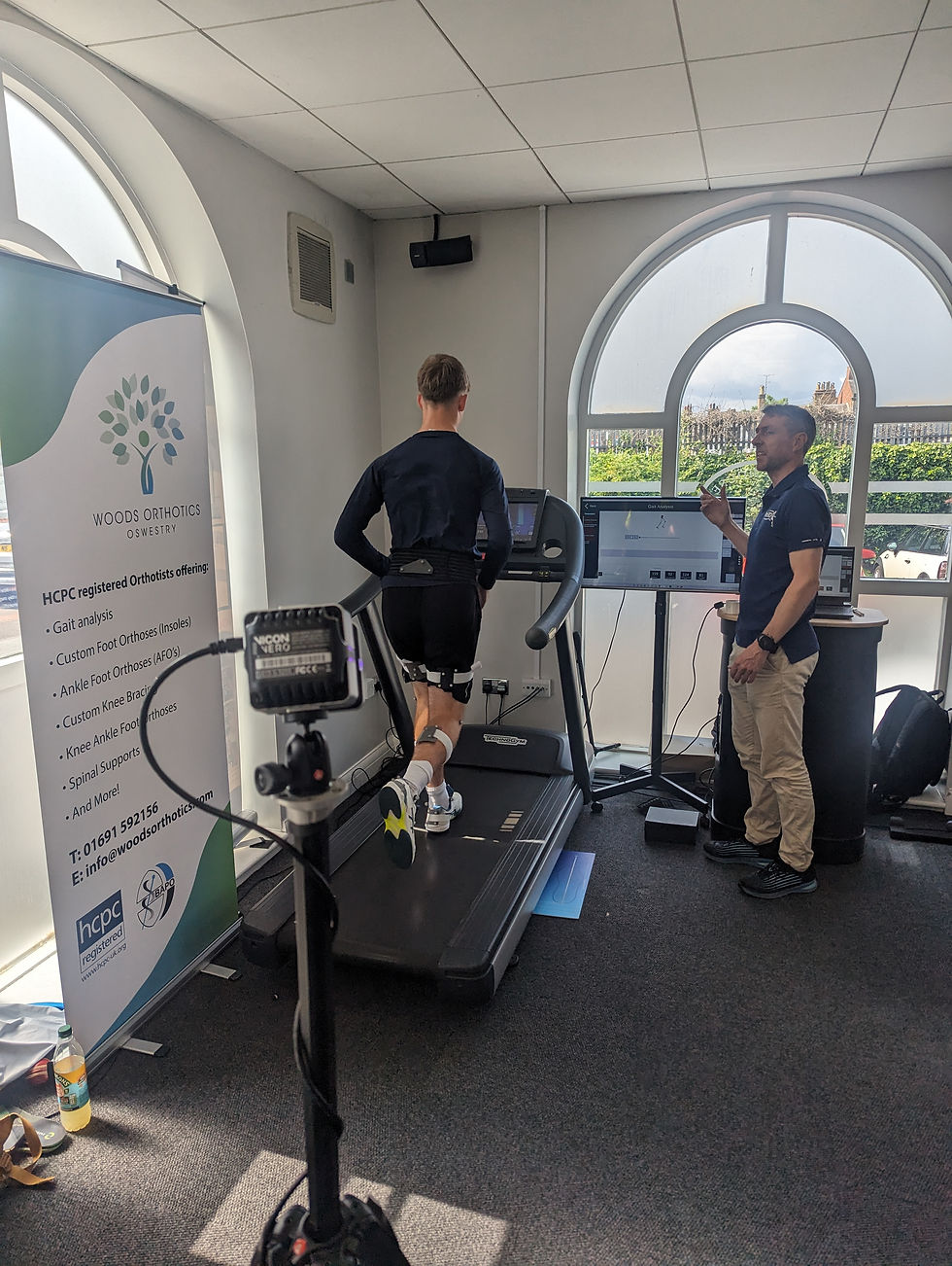 Person runs on treadmill with sensors, observed by another man. Banner reads "Woods Orthotics Oswestry." Bright room with large window.