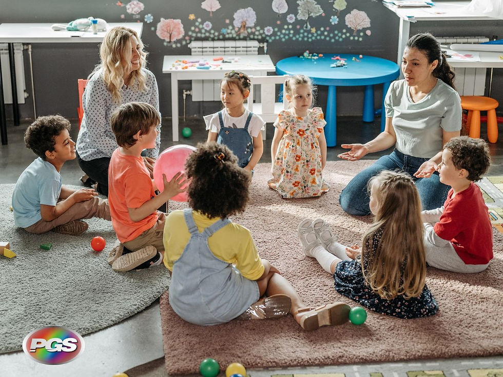 Children sit in a circle on a rug with two adults, one holding a pink ball. Classroom setting with colorful wall art and tables.