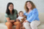 Two women and a baby sit on a couch, smiling. The baby holds a teddy bear. The setting features a white brick wall.