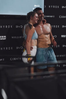 A couple at the finish line in HYROX New Zealand, smiling and posing for a photo. The woman is in white LSKD shorts and a blue crop top. The male is shirtless with blue LSKD running shorts