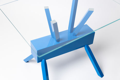 Close-up angled view of the table, emphasizing the smooth glossy blue finish of the wooden base and the clear glass top. The image captures the sleek, modern design of the table.