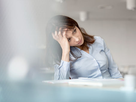 Woman in blue shirt sits at a desk, resting her head on her hand, looking thoughtful. Blurred foreground, bright setting.