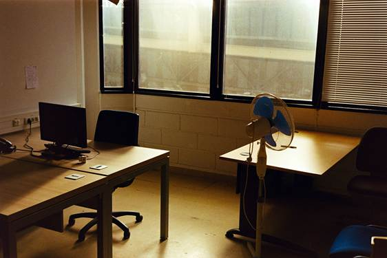 Empty office with desks, monitor, chairs, and a standing fan by sunlit windows, creating a quiet, warm atmosphere.