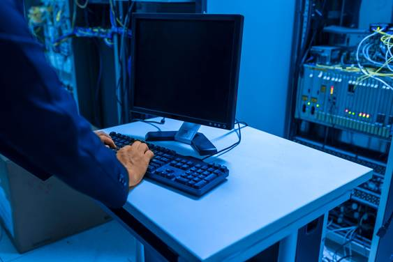 Person typing at a keyboard beside a blank monitor in a blue-lit server room with racks and cables nearby.