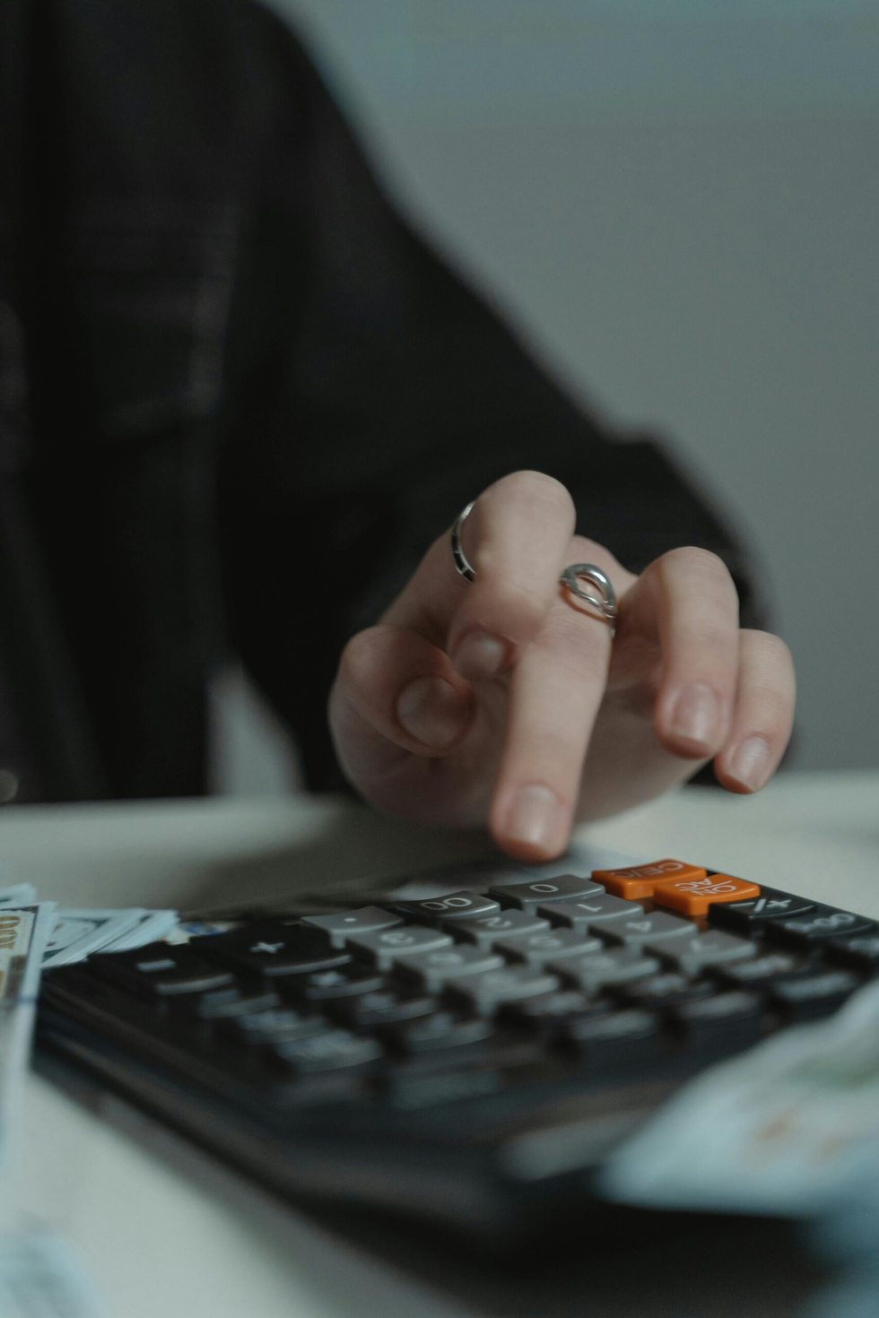 A hand with rings presses a button on a black calculator with orange keys. Blurred cash is visible, suggesting a financial context.