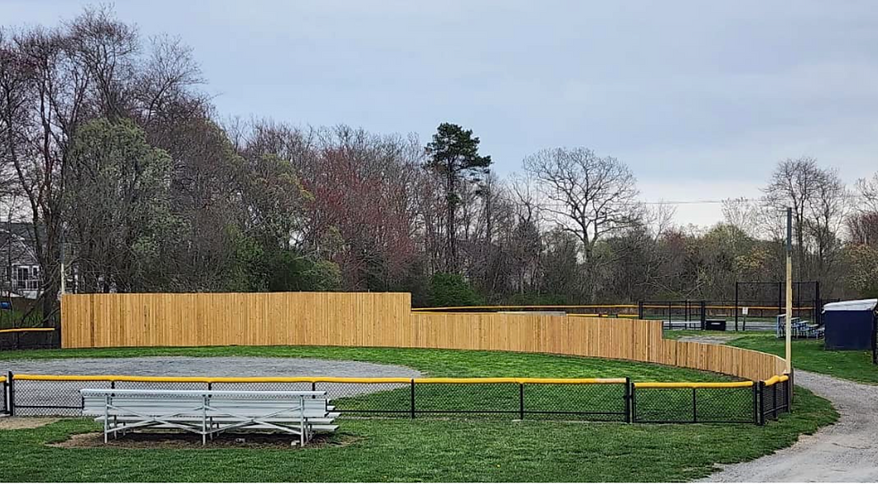 Fairhaven Baseball field with a wooden fence and bleachers. Trees line the background under a cloudy sky, creating a calm, rural setting.