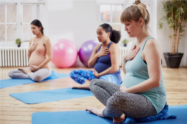 Mums are sat in easy pose in Pregnancy Yoga in Oakwood, Derby. They are sat with one hand on their bump and the other hand on their heart, with eyes closed, focusing quietly inward on their babies