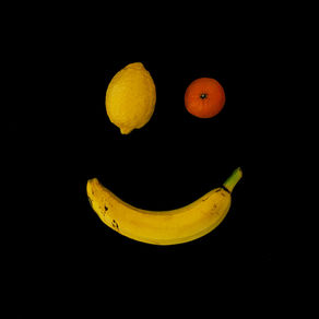 A smiley face composed of fruit: a lemon and a tangerine for eyes, and a slightly curved banana for a mouth, all set against a solid black background.