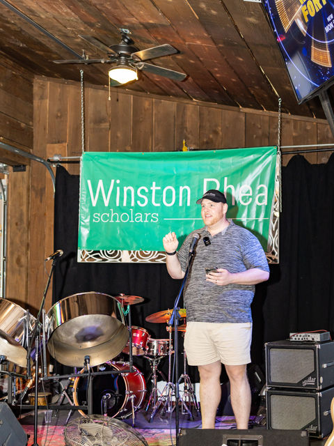 Daniel Harris, Co-Founder and Executive Director, standing on top of a stage with a Winston Rhea Banner and a set of drums in the background, giving a speech. Daniel wearing a black hat, gray polo, and khaki shorts.
