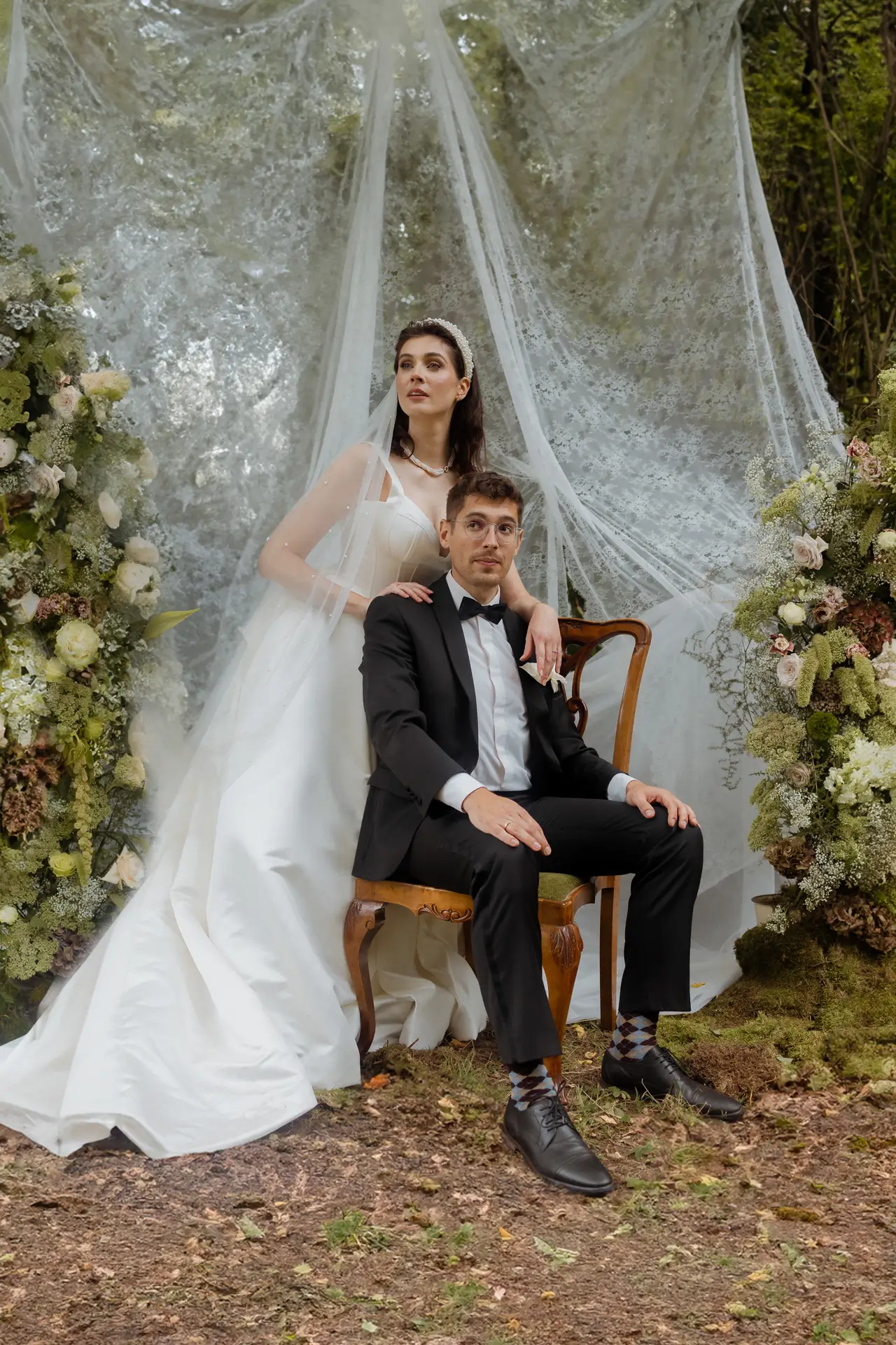 Hochzeitspaar vor Spitzenvorhang mit grüner Blumendekoration im Old Money Stil