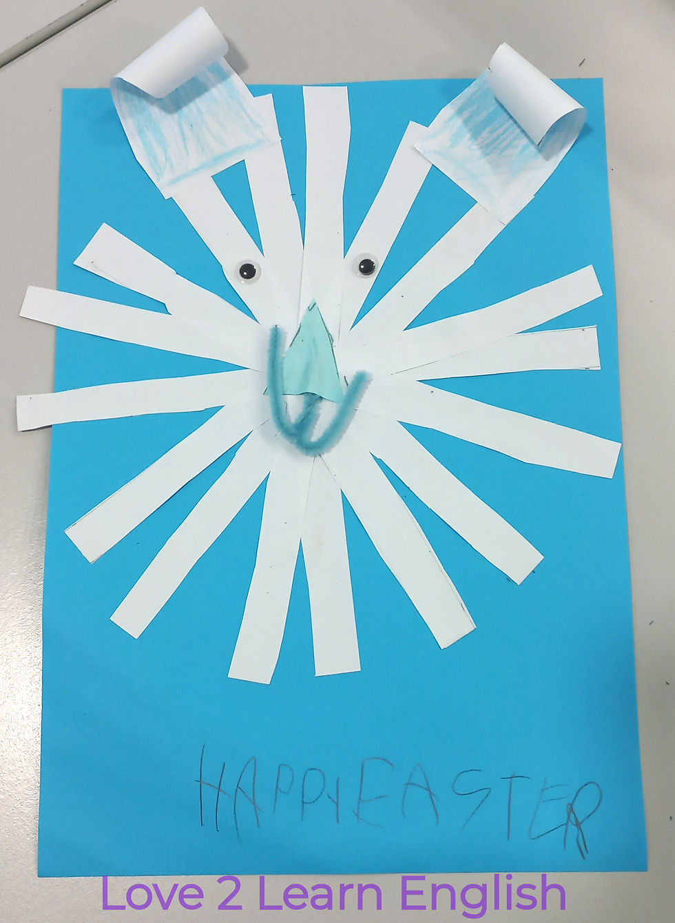 Blue paper with a white paper lion face made of strips, googly eyes, and a pipe cleaner mouth. "Happy Easter" is handwritten below.