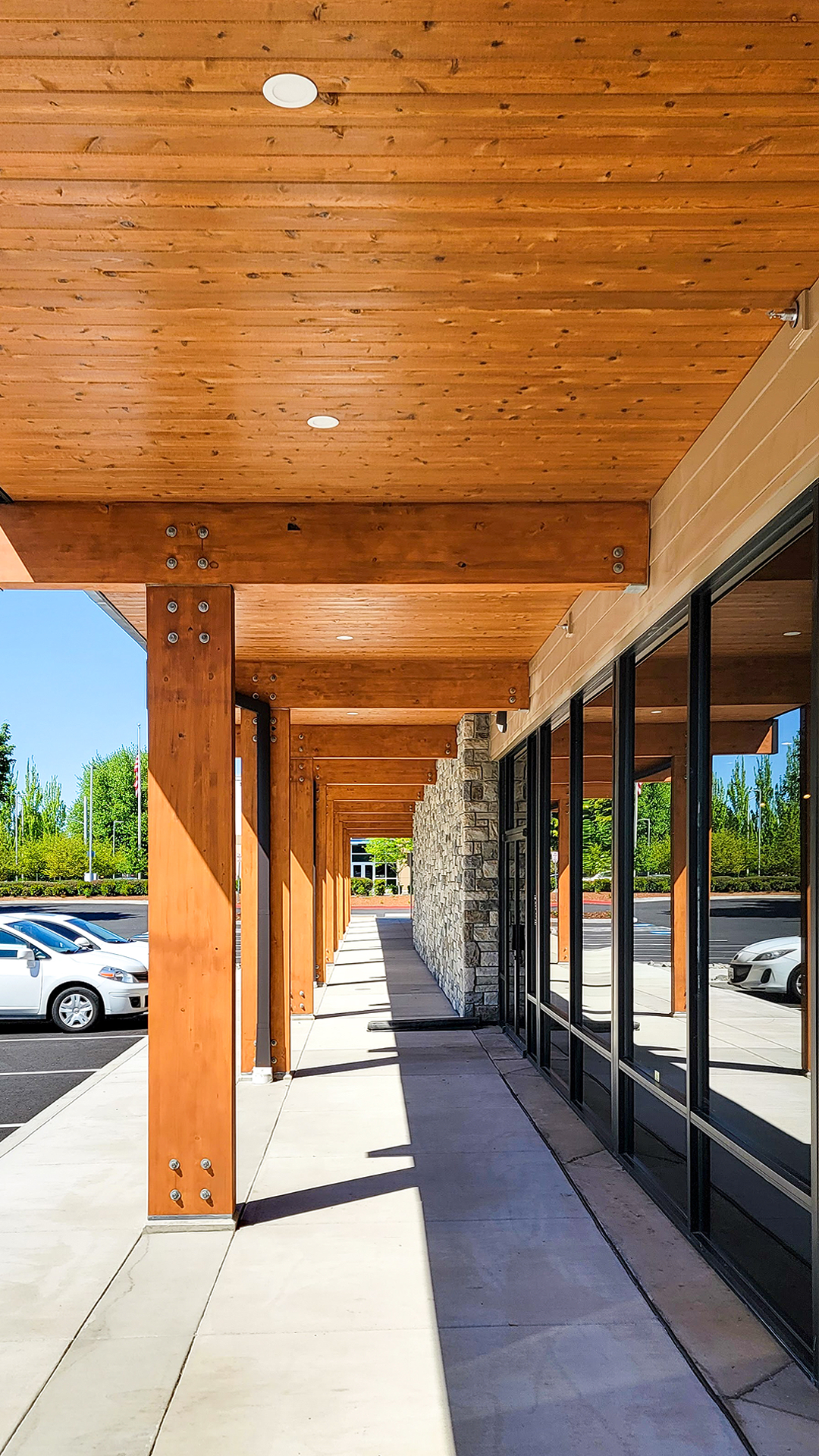 Mid day photography showing the warm wood tones of stained cedar tongue and groove soffit, knotty post and beam. To the right are black reflective storefront windows of Cascade Furniture & Mattress after commercial siding, soffit, post & beam installation by StraightLine Exteriors. 