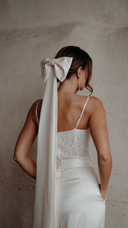 Bride wearing a sleek satin bridal bow with long tails, styled in a low bun hairstyle by Nala & Knot — a modern alternative to the traditional wedding veil by Rebecca Anne Designs.