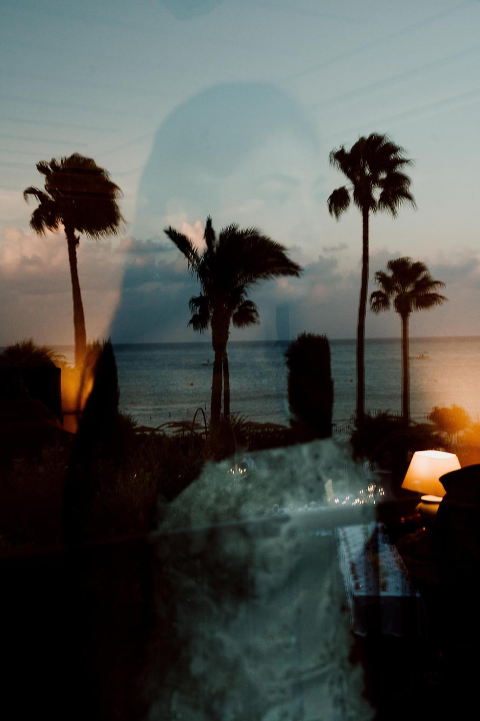 A woman's reflection on glass overlays a sunset beach scene with palm trees, creating a serene mood. A glowing lamp adds warmth.