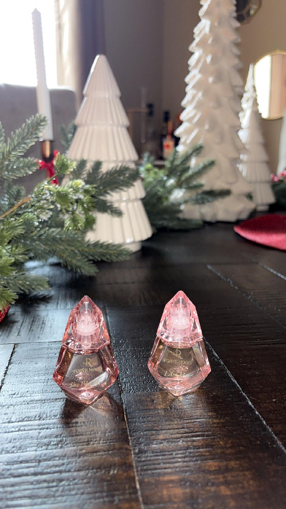 Pink perfume bottles on a dark table with white tree decorations and green foliage. Text on bottles says "I Am Edible" and "She E.O."