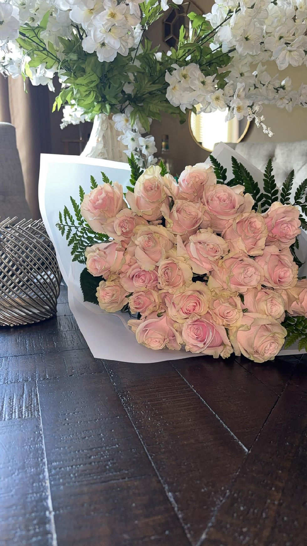 Pink rose bouquet on a dark wooden table, with white flowers behind it in a softly lit room, creating a romantic, elegant mood