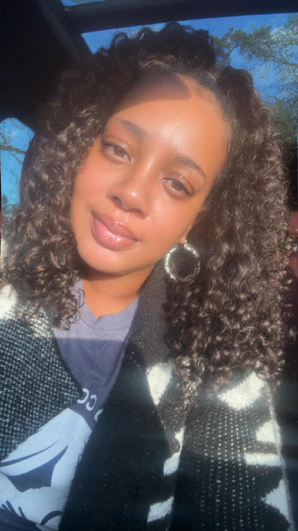Sunlit selfie of a woman with curly hair and hoop earrings in a car, looking calmly at the camera.