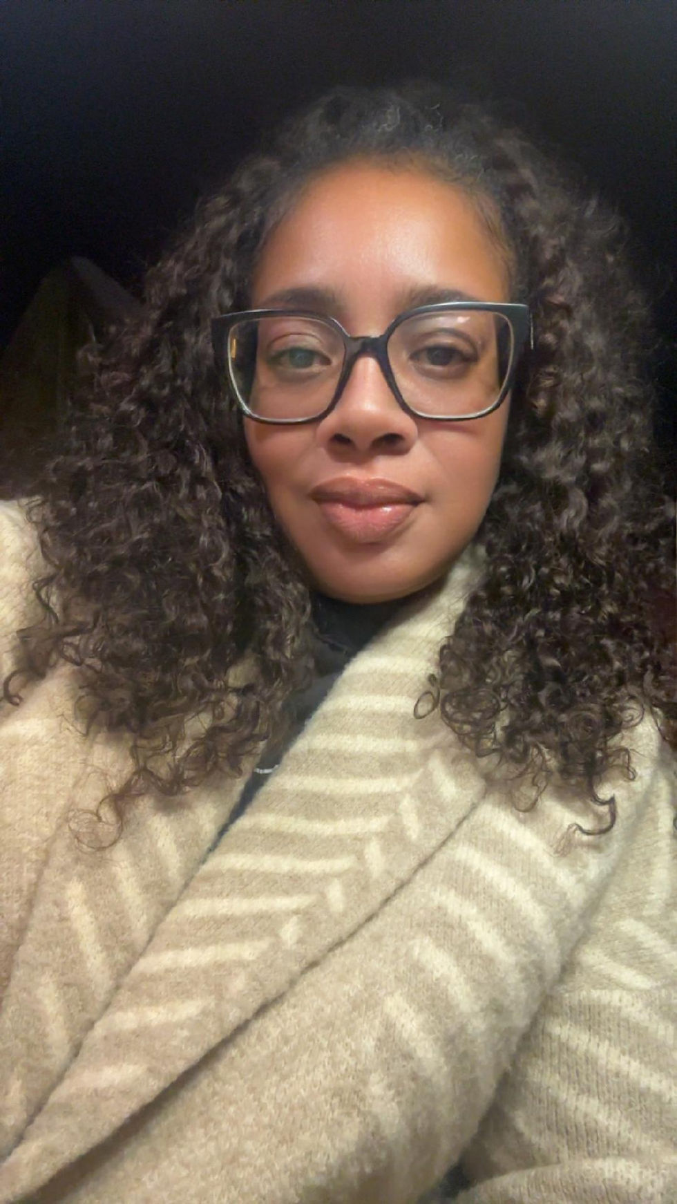 A woman with curly hair and glasses wears a beige coat with a subtle pattern. She looks content against a dark background.