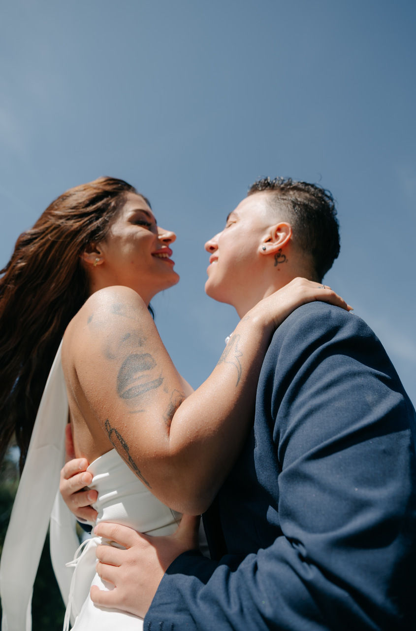 Zwei Menschen umarmen sich lächelnd vor blauem Himmel. Der eine trägt ein weißes Kleid, der andere einen blauen Anzug. Tattoos sichtbar.