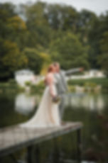 Ein Brautpaar steht auf einem Steg vor einem See. Der Mann zeigt in die Ferne. Sie trägt Blumen. Hintergrund: grüne Bäume, Gebäude.