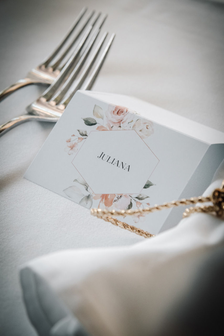 Tischkarte mit Blumenmuster und dem Namen Juliana auf weißer Tischdecke, daneben zwei silberne Gabeln und goldene Schnur.