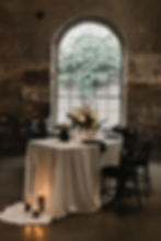 Ein elegant gedeckter Tisch mit weißen Blumen, Kerzen und schwarzen Stühlen vor einem großen Bogenfenster in einem rustikalen Raum.