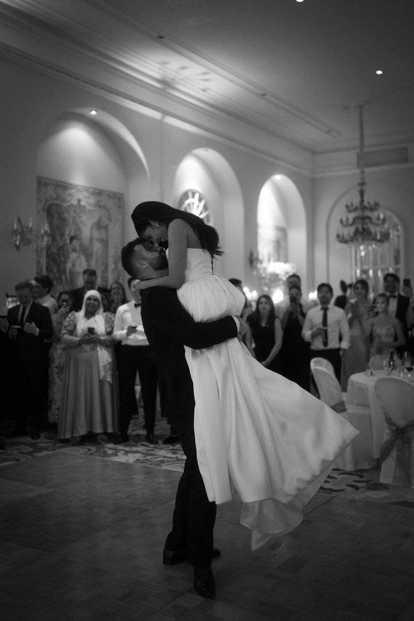 Ein Brautpaar tanzt eng umschlungen in einem eleganten Saal, umgeben von applaudierenden Gästen. Die Stimmung ist festlich und romantisch.