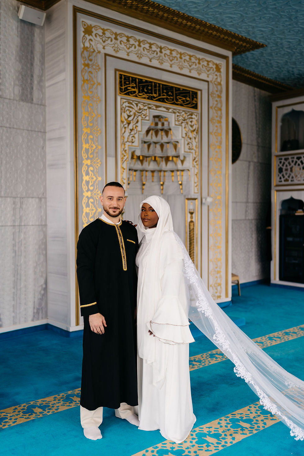 Brautpaar in Schwarz und Weiß in prunkvoller Moschee mit blauem Teppich und Goldornamenten, ruhig posierend.