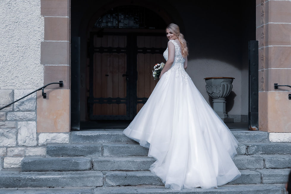 Braut in weißem Kleid steht auf einer Steintreppe vor einer Holztür, hält Blumenstrauß und lächelt zurück. Eleganter, freudiger Moment.