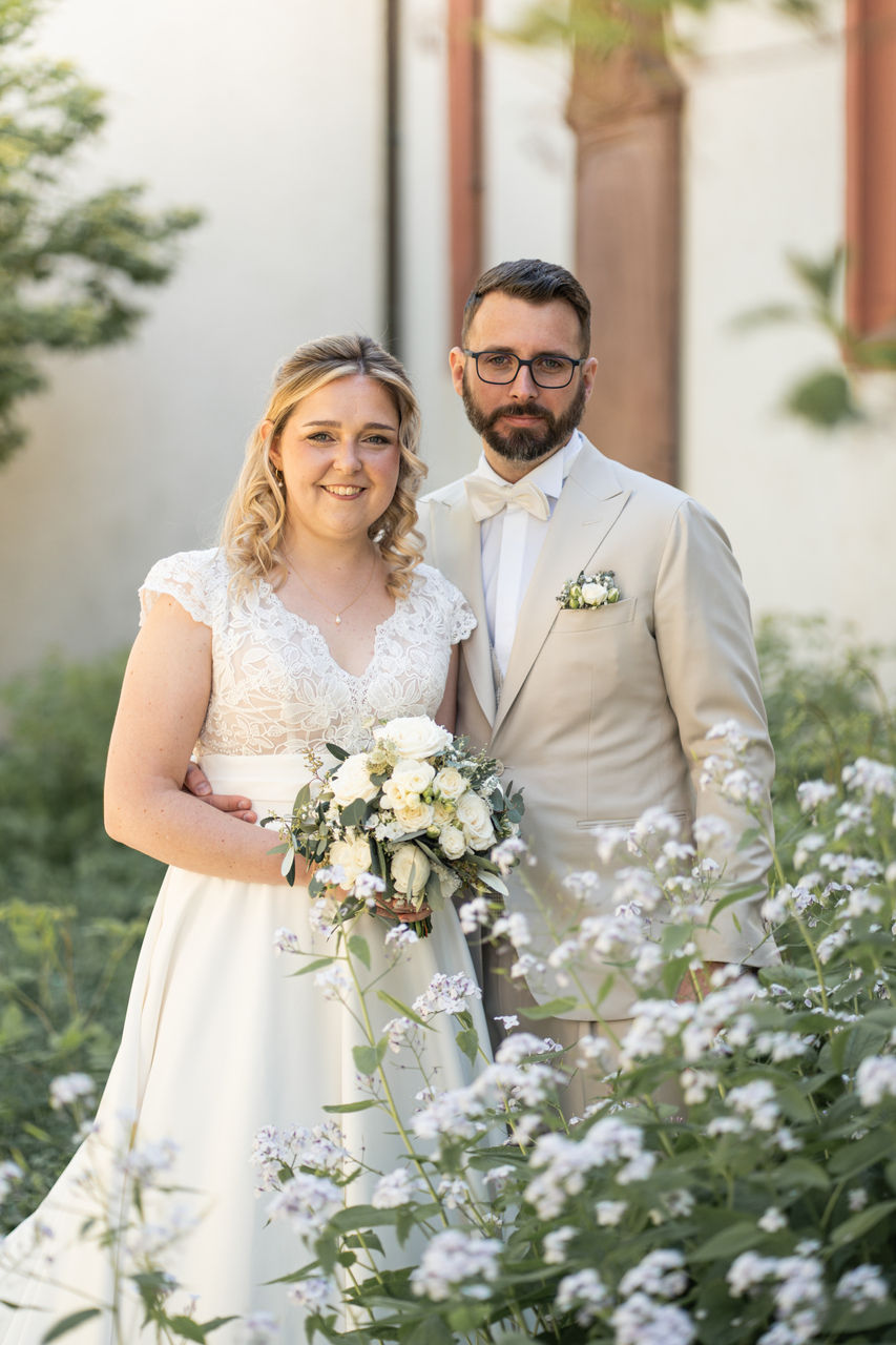 Brautpaar in Weiß im Garten: lächelnde Braut mit Blumenstrauß, Bräutigam im hellen Anzug, Blumen im Vordergrund.