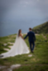 Brautpaar in Hochzeitskleidung hält Hände in den Bergen. Die Braut in Weiß, der Bräutigam in Blau. Wolkiger Himmel, romantische Stimmung.
