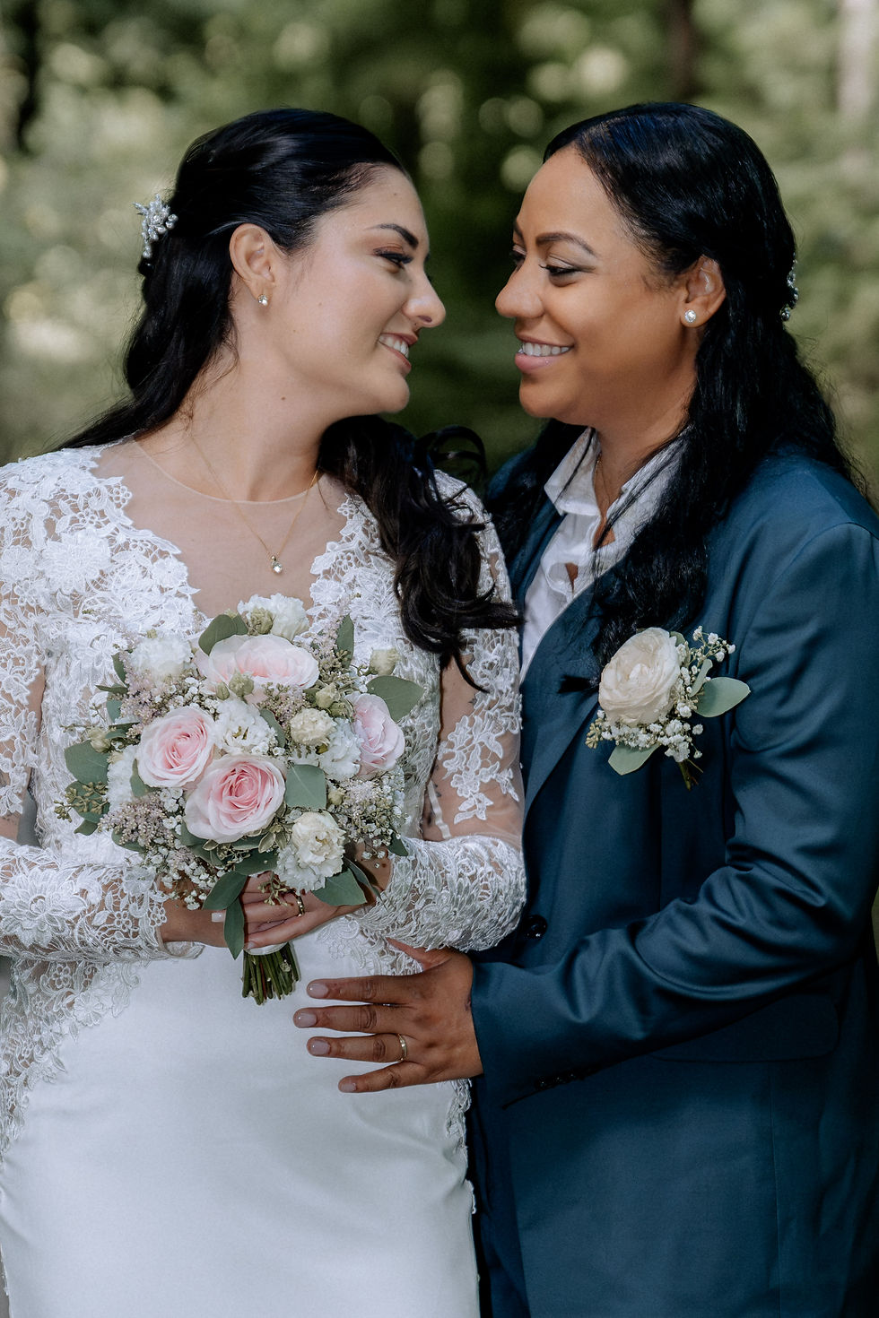 Zwei lächelnde Frauen in Hochzeitskleidung stehen im Grünen, halten sich mit weißen und rosa Blumensträußen eng umarmt. Glücklich und verliebt.