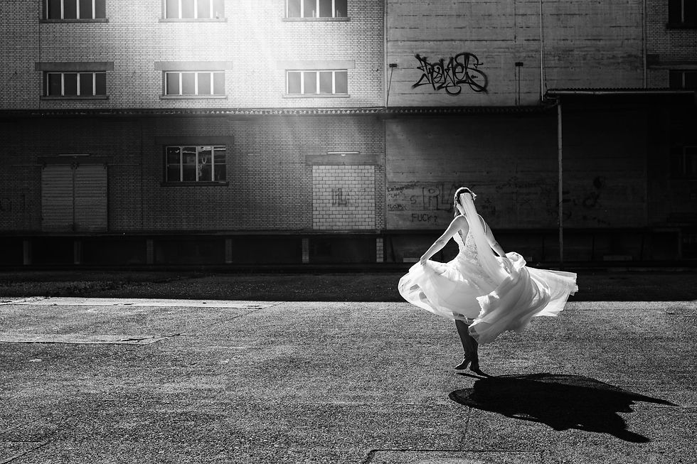 Braut tanzt in weißem Kleid auf leerer Straße vor industrieller Backsteinfassade mit Graffiti. Sonnenstrahlen erzeugen dramatisches Licht.