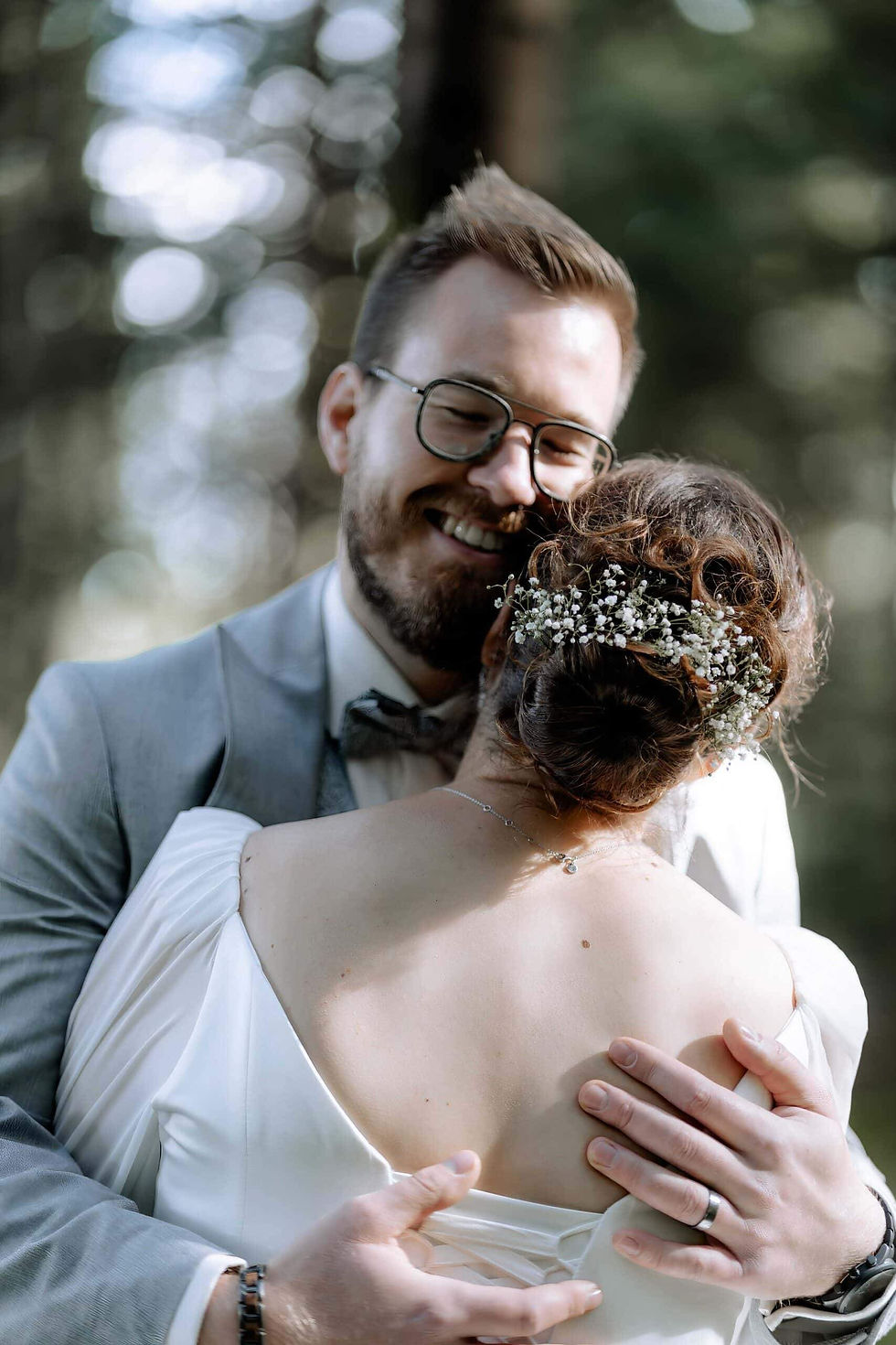 Paar umarmt sich lächelnd im Wald. Er trägt einen grauen Anzug, sie ein weißes Kleid mit Blumen im Haar. Sonnenlicht fällt durch Bäume.