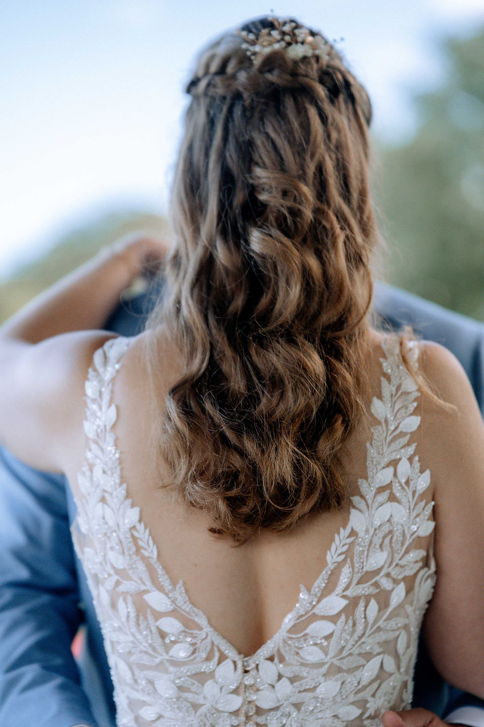 Braut mit lockigem Haar in Spitze verziertem Kleid umarmt Partner im Freien. Unscharfer grüner Hintergrund, romantische Stimmung.