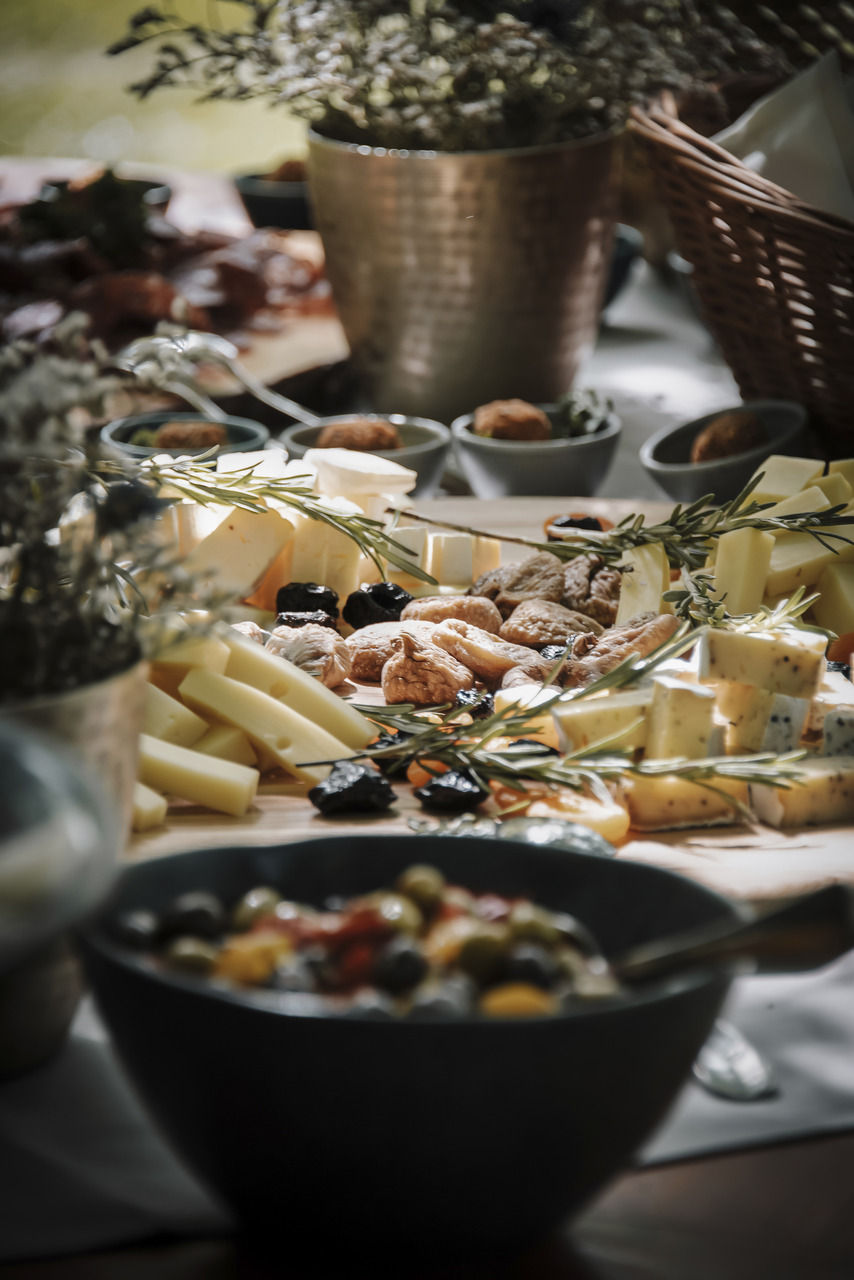 Eine Vielfalt an Käsescheiben und getrockneten Früchten mit Kräutern auf einem Brett, umgeben von Schalen und einem Korb in natürlichem Licht.