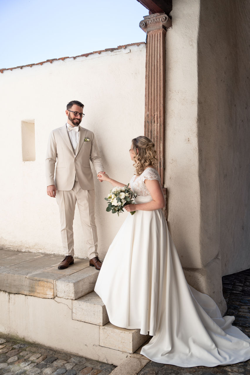 Braut im weißen Kleid mit Blumenstrauß reicht dem Bräutigam im beigen Anzug die Hand vor heller Mauer.