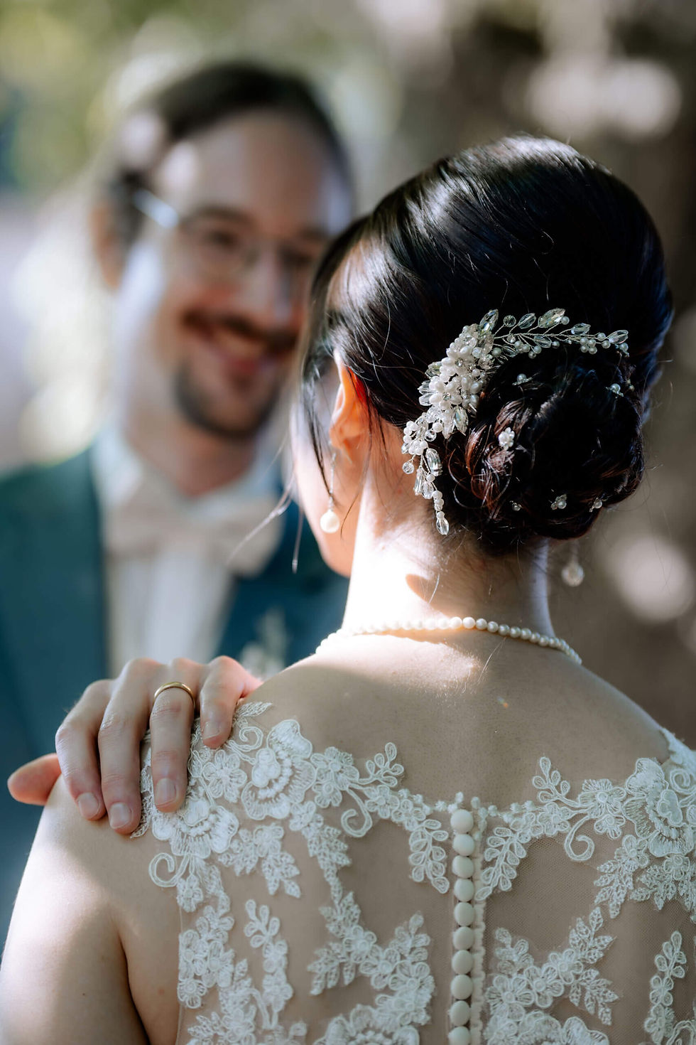 Brautpaar lächelt sich an. Die Braut trägt ein Spitzenkleid und Haaraccessoire, während er ihre Schulter hält. Sanfte, romantische Stimmung.