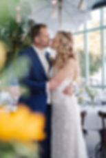 Brautpaar küsst sich unter weißen Regenschirmen in elegant dekoriertem Saal; Bräutigam in blauem Anzug, Braut im Spitzenkleid.