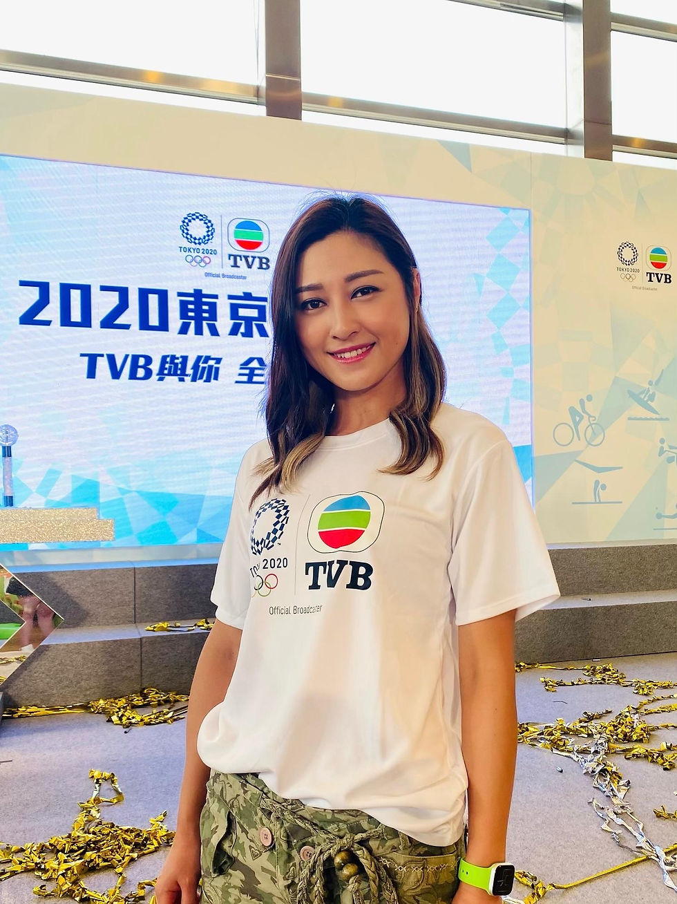 李思雅身穿白色TVB奧運T恤與迷彩長褲,站在2020東京奧運直播佈景前,地上散落金色紙碎