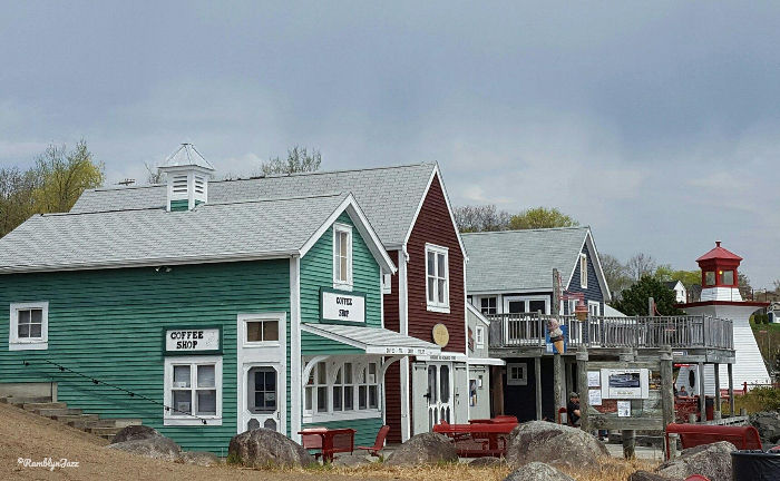 Colourful buildings line a street, with a green coffee shop in front. Overcast sky and lighthouse in the background create a quaint scene.