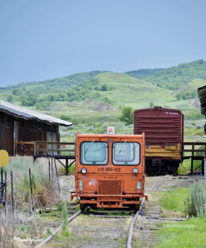 Orange railcar on tracks with wooden buildings and cargo train in background, set in lush green hills under a cloudy sky.