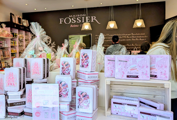 Maison Fossier shop with pastel gift tins and wrapped baskets on display; customers browse under hanging lights.