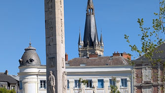 Stone war memorial in a sunny town square, with a church spire and blue-shuttered buildings under a clear blue sky.