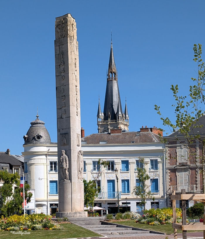 Stone war memorial in a sunny town square, with a church spire and blue-shuttered buildings under a clear blue sky.