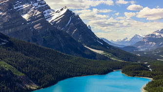 Majestic mountain range with snowcapped peaks overlooks a vibrant turquoise lake. Lush forest surrounds the serene, picturesque landscape.