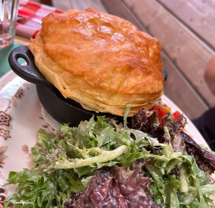 Golden pastry-topped dish in a black pot with fresh green salad on a patterned plate. Wooden background creates a cozy atmosphere.