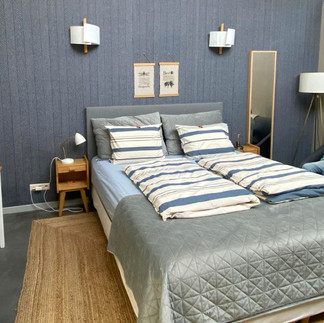Cozy bedroom with a gray quilted bed, striped pillows, blue walls, and warm lighting. Two framed prints on the wall add charm.